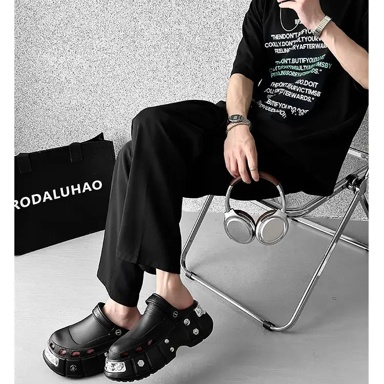 A person seated on a folding chair wearing a black graphic T-shirt, black pants, and chunky black platform clogs with silver studs and clear embellishments. A black cap, silver headphones, and a black RODALUHAO tote bag are also visible against a textured gray wall.