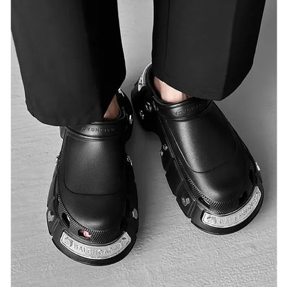 A person wearing black chunky platform clogs with silver metal accents, paired with black trousers, standing on a gray concrete floor. White text is overlaid on the upper left.