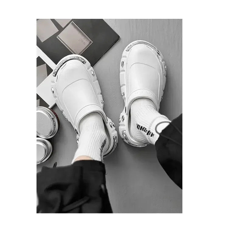 A person wearing white slip-on clog-style shoes with chunky sculpted soles and white socks, photographed from above on a gray floor with a magazine and small round objects nearby.