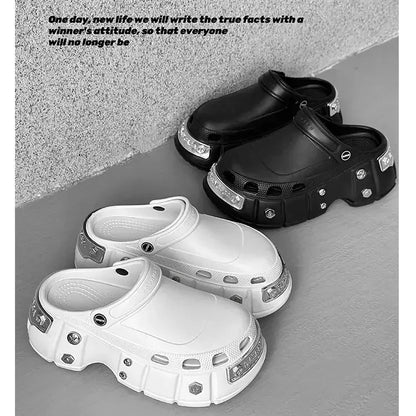 Two pairs of chunky clog-style shoes shown on a gray floor against a textured gray wall. The foreground pair is white with silver metallic studs and clear rhinestone-look trim, and the background pair is black with matching silver accents.