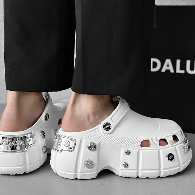 White chunky platform clogs with silver rhinestone embellishments, shown on a model wearing black pants. A black bag with the partial brand name 'DALU' is visible in the background.