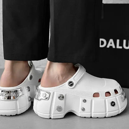 White chunky platform clogs with silver rhinestone embellishments, shown on a model wearing black pants. A black bag with the partial brand name 'DALU' is visible in the background.