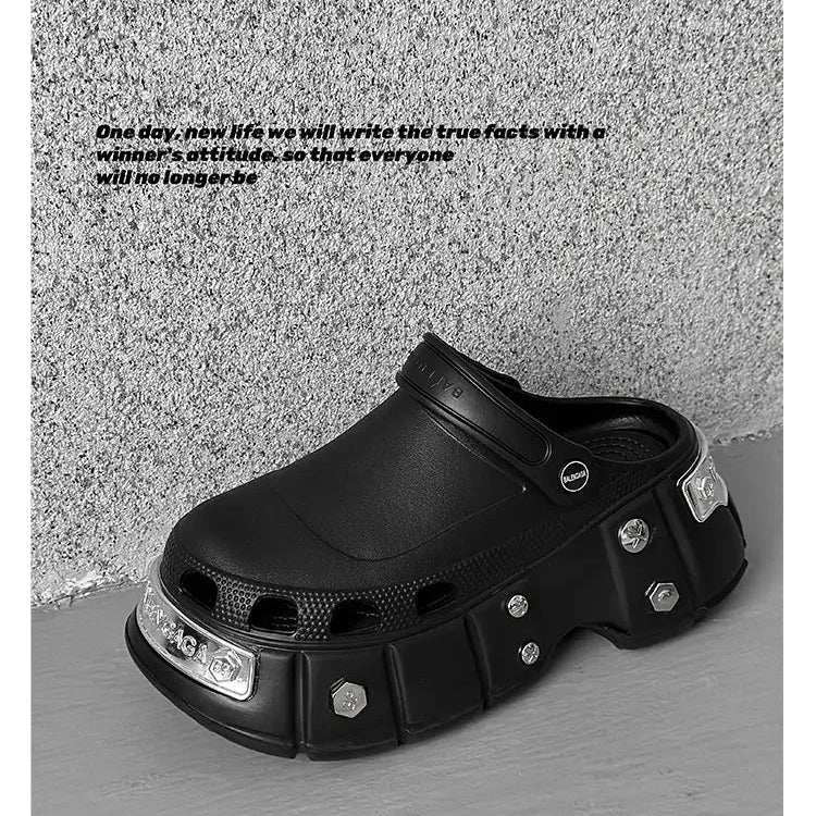 A black chunky clog-style shoe with metallic silver hexagon accents and a textured sole, photographed against a gray concrete-textured wall and floor. Black text is printed on the wall to the left.