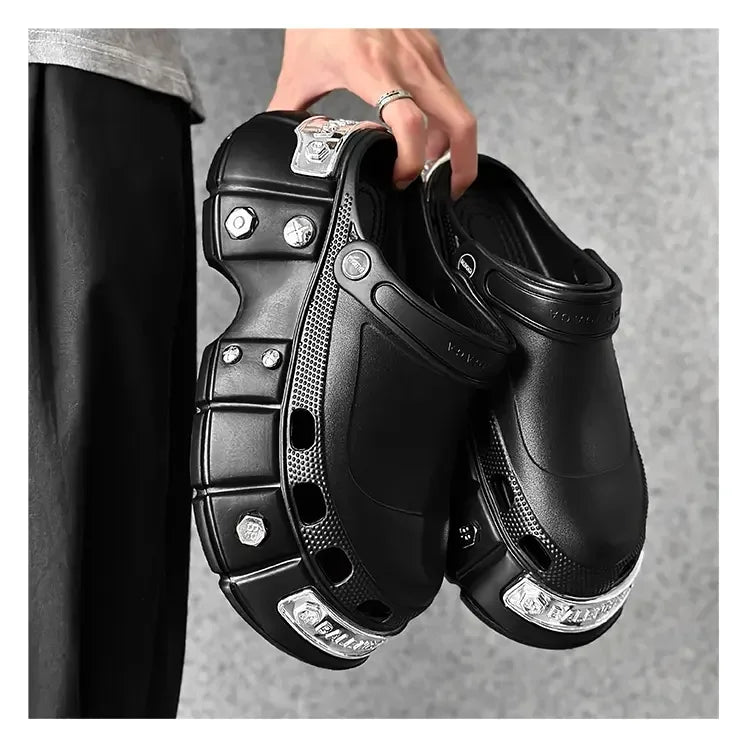 A person holds a pair of chunky black clog-style shoes with metallic silver decorative accents and textured paneling against a gray background.
