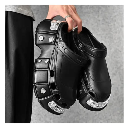 A person holds a pair of chunky black clog-style shoes with metallic silver decorative accents and textured paneling against a gray background.