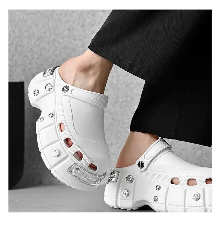 A pair of chunky white clog-style mule shoes with silver metal studs and hardware, shown being worn with black pants against a gray studio background.