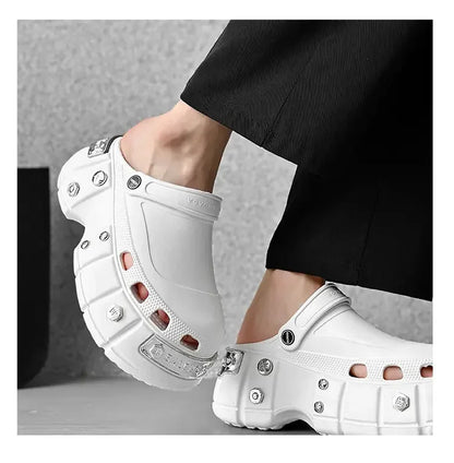 A pair of chunky white clog-style mule shoes with silver metal studs and hardware, shown being worn with black pants against a gray studio background.