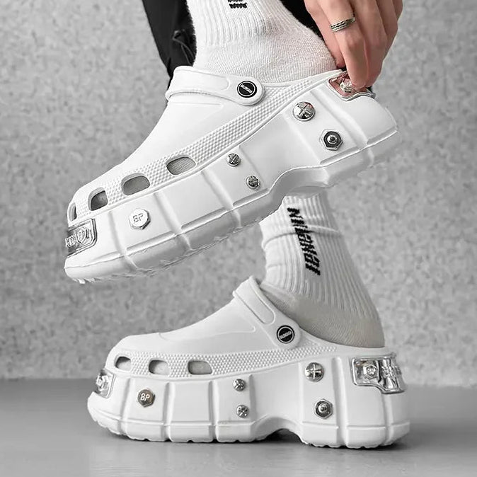 A person wearing chunky white platform clog-style shoes with metal studs and clear rhinestone embellishments, paired with white crew socks printed with the brand name 'Meganol'.