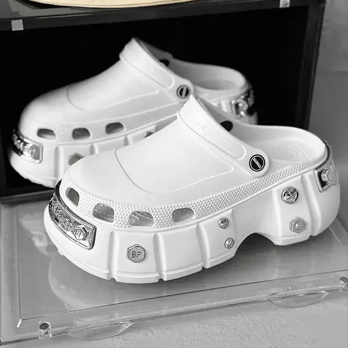 A pair of chunky white platform clogs with silver metal studs and hardware, displayed on a clear acrylic stand next to a black box. A beige bucket hat with a black smiley face graphic and the word 'Smile' is resting on top of the box.