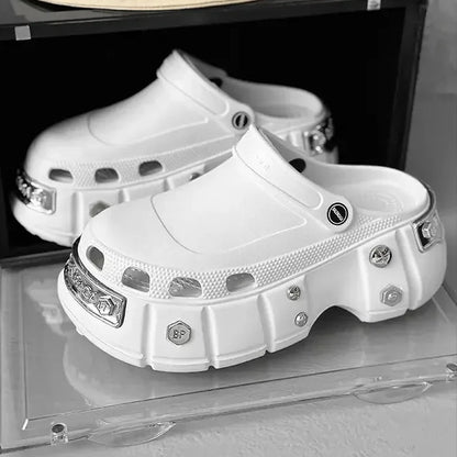 A pair of chunky white platform clogs with silver metal studs and hardware, displayed on a clear acrylic stand next to a black box. A beige bucket hat with a black smiley face graphic and the word 'Smile' is resting on top of the box.