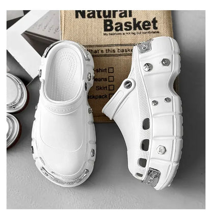 A pair of chunky white platform clogs with silver metal rivet details, displayed on a gray surface with a cardboard box in the background.