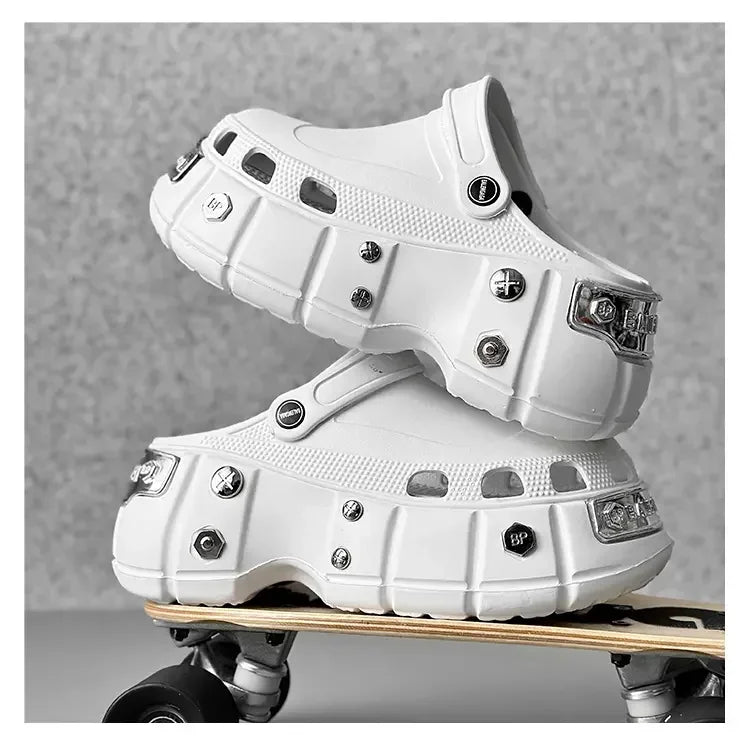 A pair of chunky white clog-style shoes with metallic hardware, stacked on a skateboard against a textured gray background.