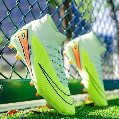 Green soccer cleats with orange accents on grass near a chain-link fence.