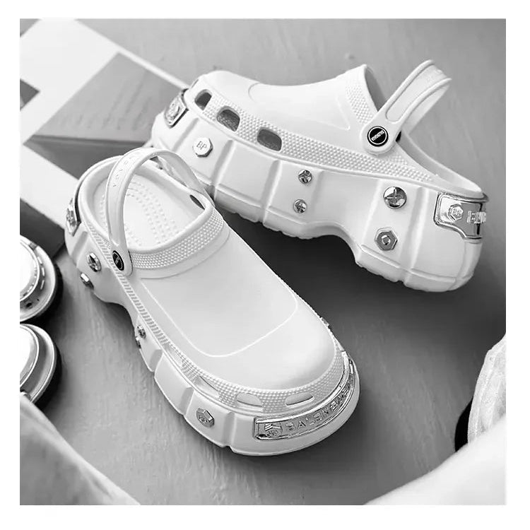 A pair of chunky white clog-style shoes with metallic studs and rivet details, displayed on a neutral gray surface.