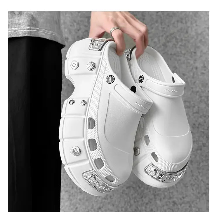 A person holds a pair of chunky white clog-style shoes with decorative silver metal studs and embellishments. The background is a textured gray wall.