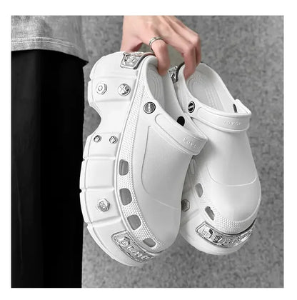 A person holds a pair of chunky white clog-style shoes with decorative silver metal studs and embellishments. The background is a textured gray wall.
