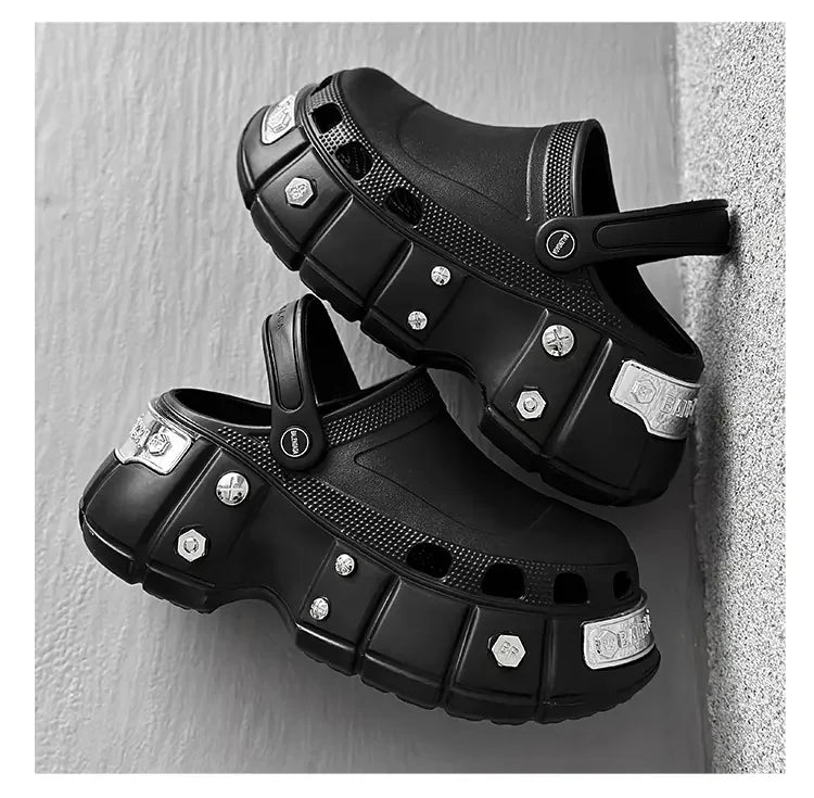 A pair of black clog-style sandals with chunky platform soles and decorative silver-tone studs, shown against a light gray textured wall.