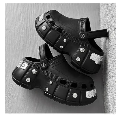 A pair of black clog-style sandals with chunky platform soles and decorative silver-tone studs, shown against a light gray textured wall.